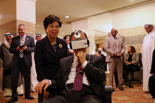 UN Secretary-General Ban Ki-moon sitting in a chair with a VR headset on, watching Clouds over Sidra, Executive Director, WHO Margaret Chan is standing by his side and they are both surrounded by Kuwaiti dignitaries.
