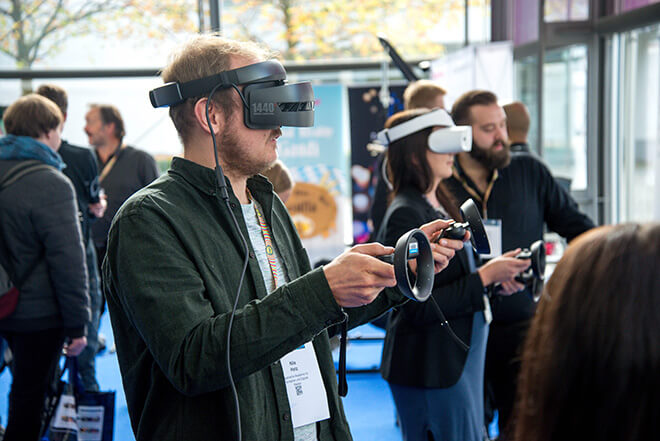 Man going through a VR demo, standing with a headset on, surrounded by other people trying and some other facilitating the demo.